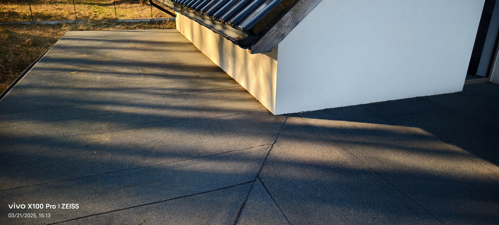 Płaski dach pokryty czarną papą termozgrzewalną, widoczne łączenia i cień rynny na jasnej ścianie budynku.