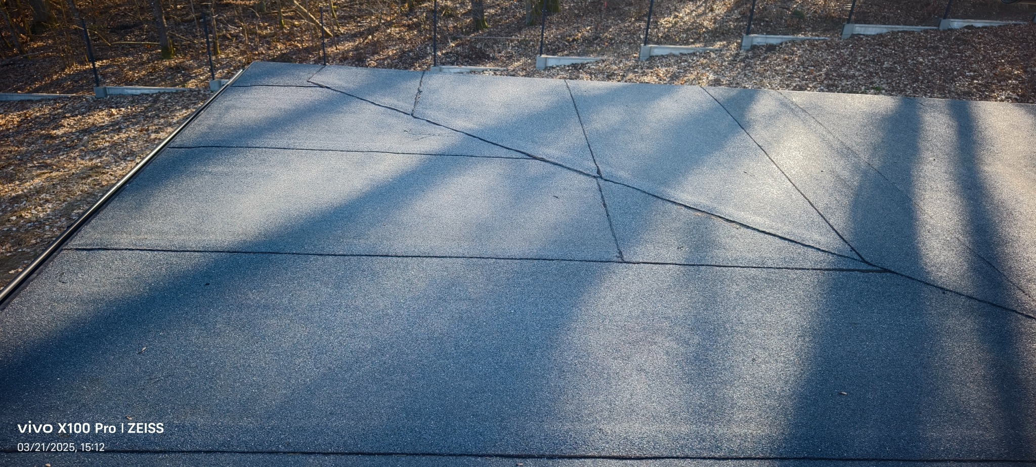 Płaski dach pokryty papą termozgrzewalną, widoczne łączenia i cień drzewa, w tle drzewa liściaste.