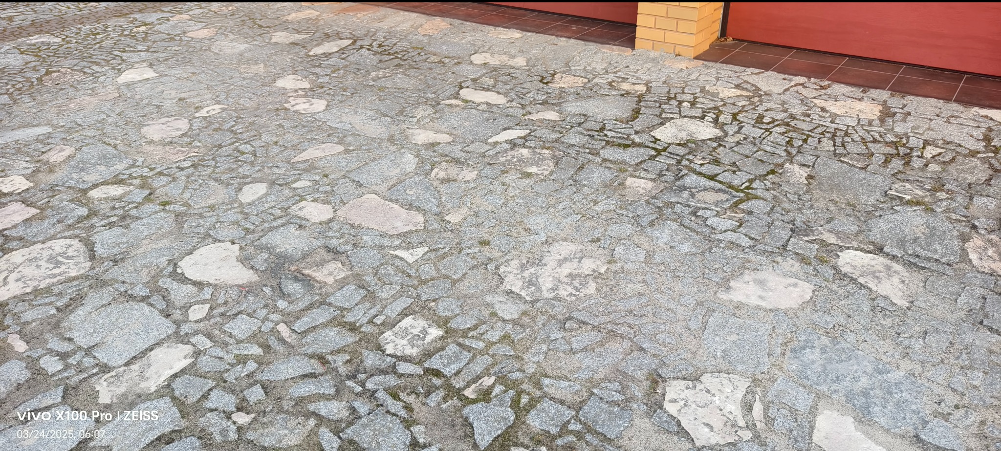 Szara kostka brukowa o nieregularnym kształcie i wielkości, ułożona przed budynkiem z widocznym fragmentem bramy garażowej i ceglanego narożnika.