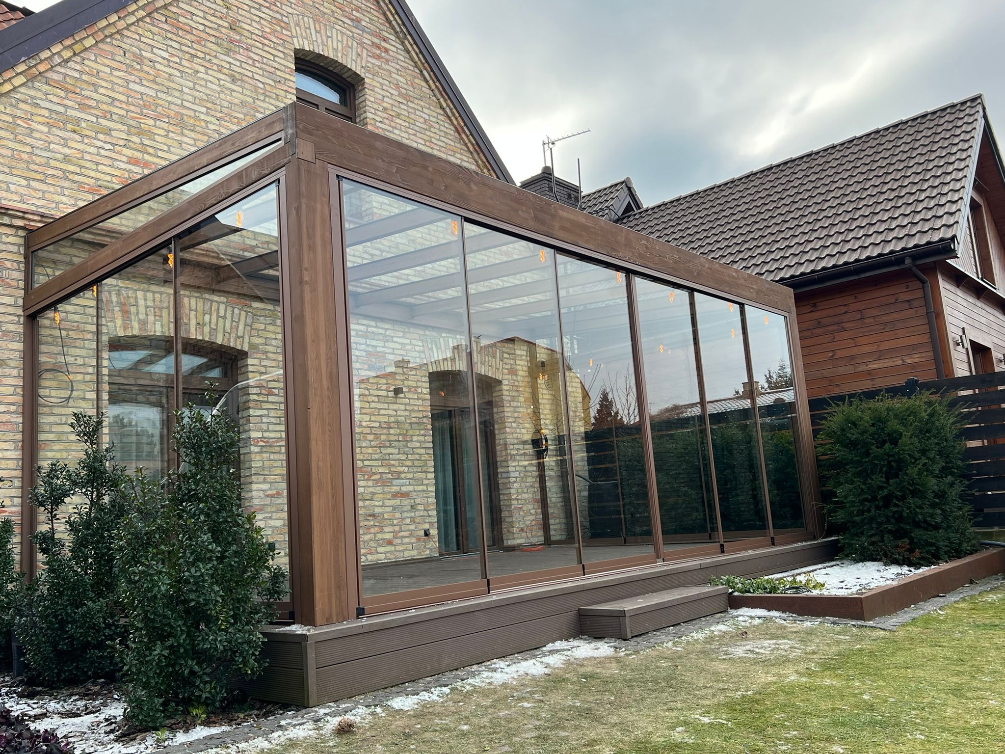 Zadaszony taras z brązową konstrukcją i szklanymi ścianami przylegający do ceglanego domu, widok z zewnątrz na trawnik z resztkami śniegu.