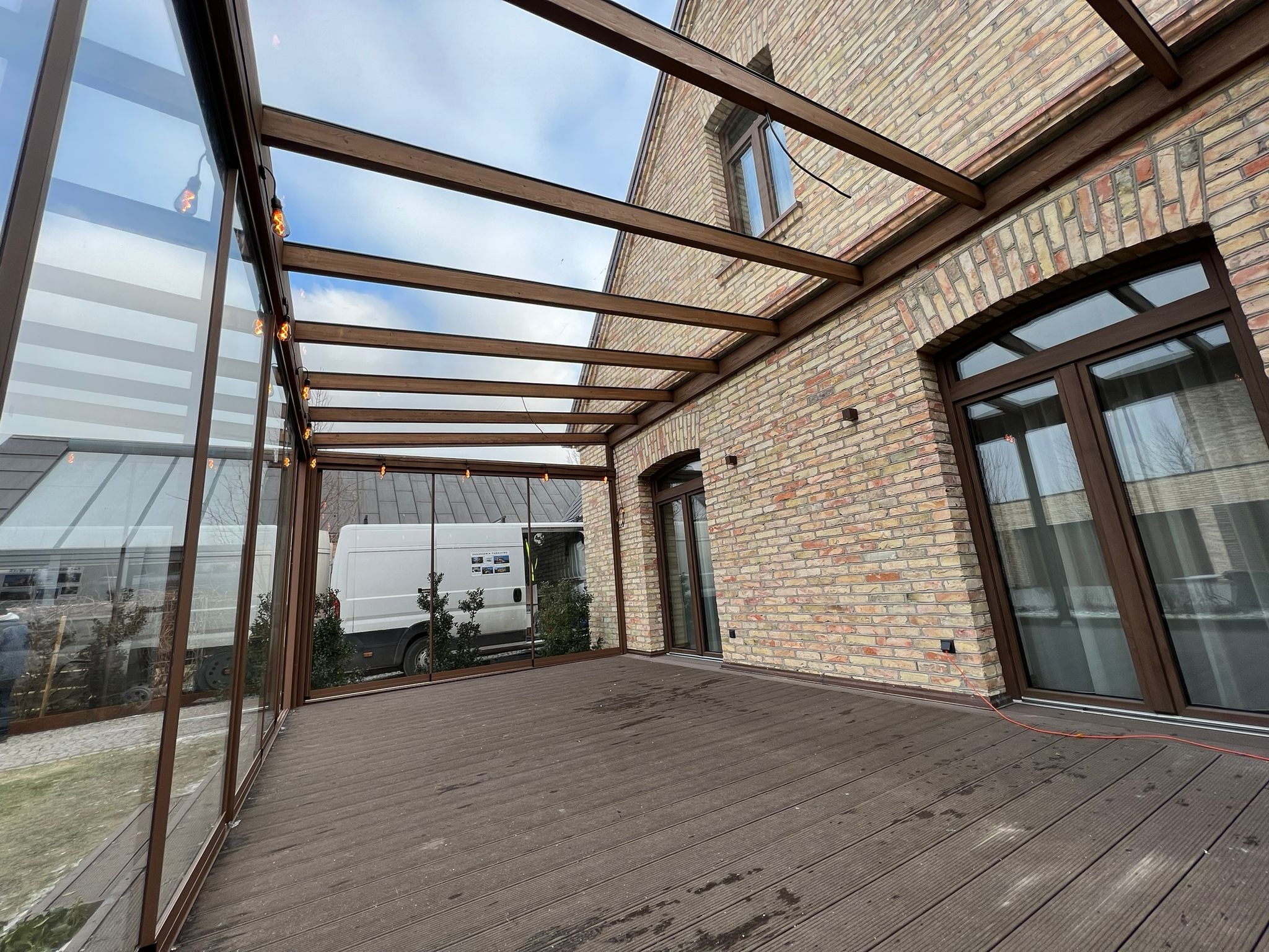 Zadaszony taras z kompozytowych desek, przeszklonymi ścianami i drewnianą konstrukcją nośną, oświetlony girlandą żarówek, z widokiem na ceglany budynek i zaparkowany samochód dostawczy.