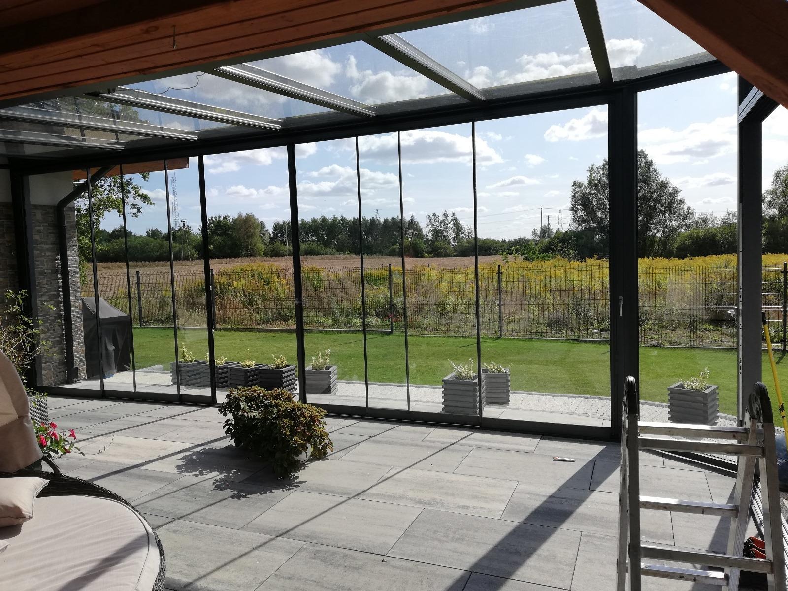 Zadaszony taras z przesuwnymi szklanymi panelami, widok na ogród i pole, szare płytki tarasowe, rośliny w betonowych donicach, drabina w rogu.