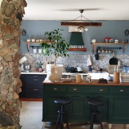 F.U.H Geppetto Łukasz Szefeciński - Kuchnia w stylu rustykalnym z kamienną kolumną, drewnianymi belkami i zieloną wyspą. Niebieskie ściany, mozaika nad blatem i drewniana podłoga tworzą przytulny klimat.