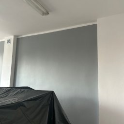 F.U.H Geppetto Łukasz Szefeciński - Ściana pomalowana na szaro, zabezpieczona czarną folią malarską. Widoczny fragment sufitu z lampą. Ujęcie z dołu, prezentujące etap prac malarskich.