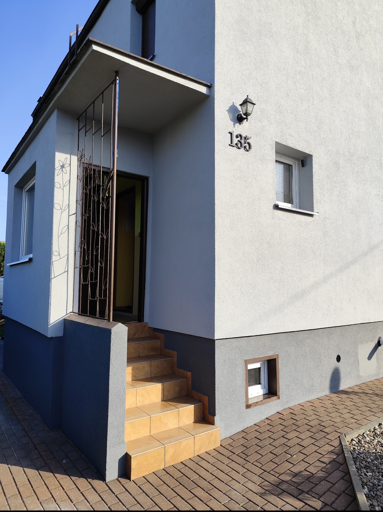 Elewacja domu z numerem 135, schodami wejściowymi wyłożonymi płytkami i ozdobną kratą przy wejściu. Widoczny fragment chodnika z kostki brukowej.