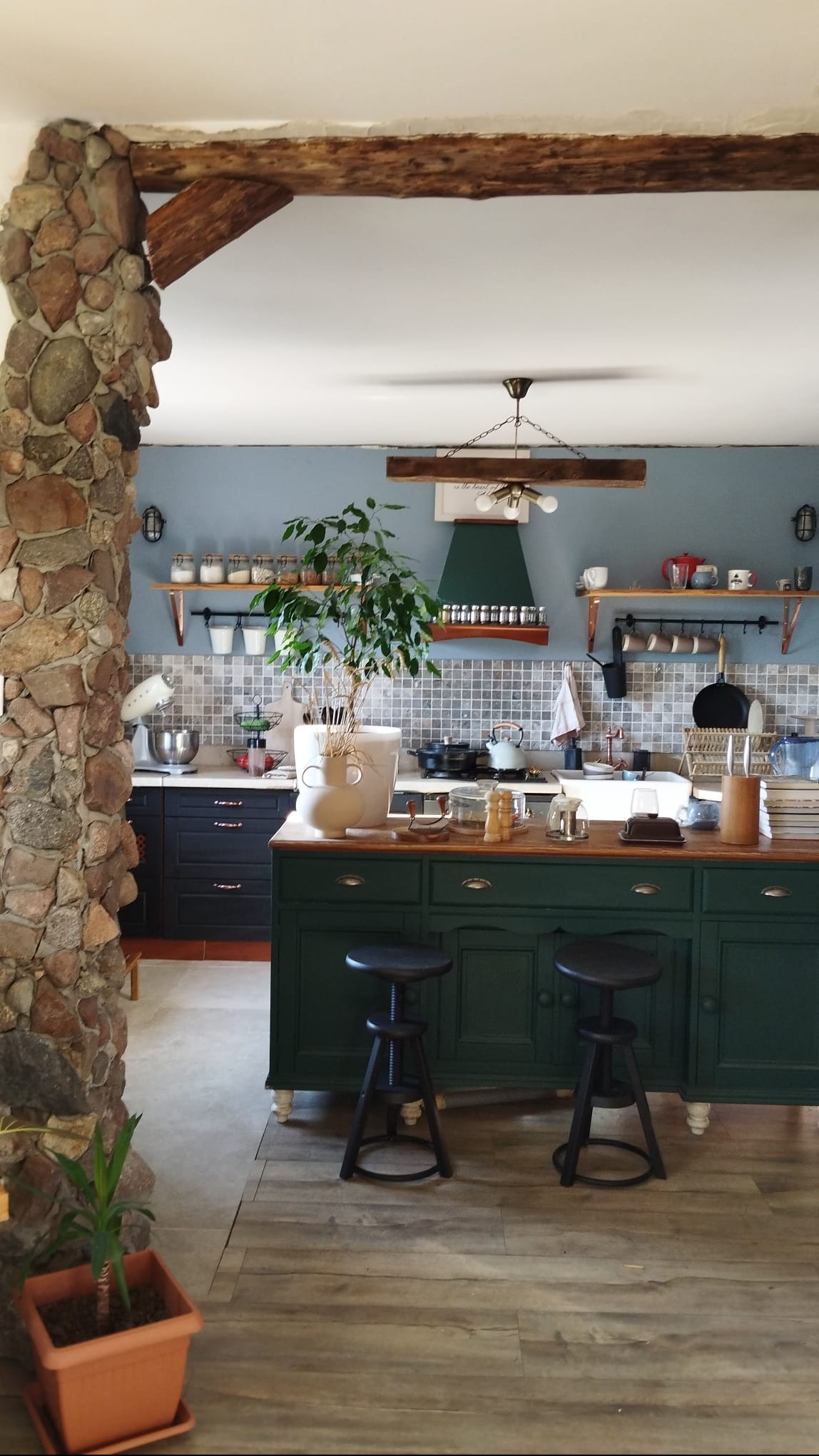 Kuchnia w stylu rustykalnym z kamienną kolumną, drewnianymi belkami i zieloną wyspą. Niebieskie ściany, mozaika nad blatem i drewniana podłoga tworzą przytulny klimat.