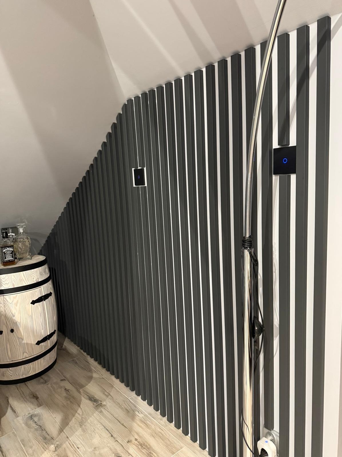 Ściana z pionowych, szarych listew w skosie poddasza, z dwoma czarnymi włącznikami dotykowymi. Obok drewniana beczka i srebrna lampa podłogowa.