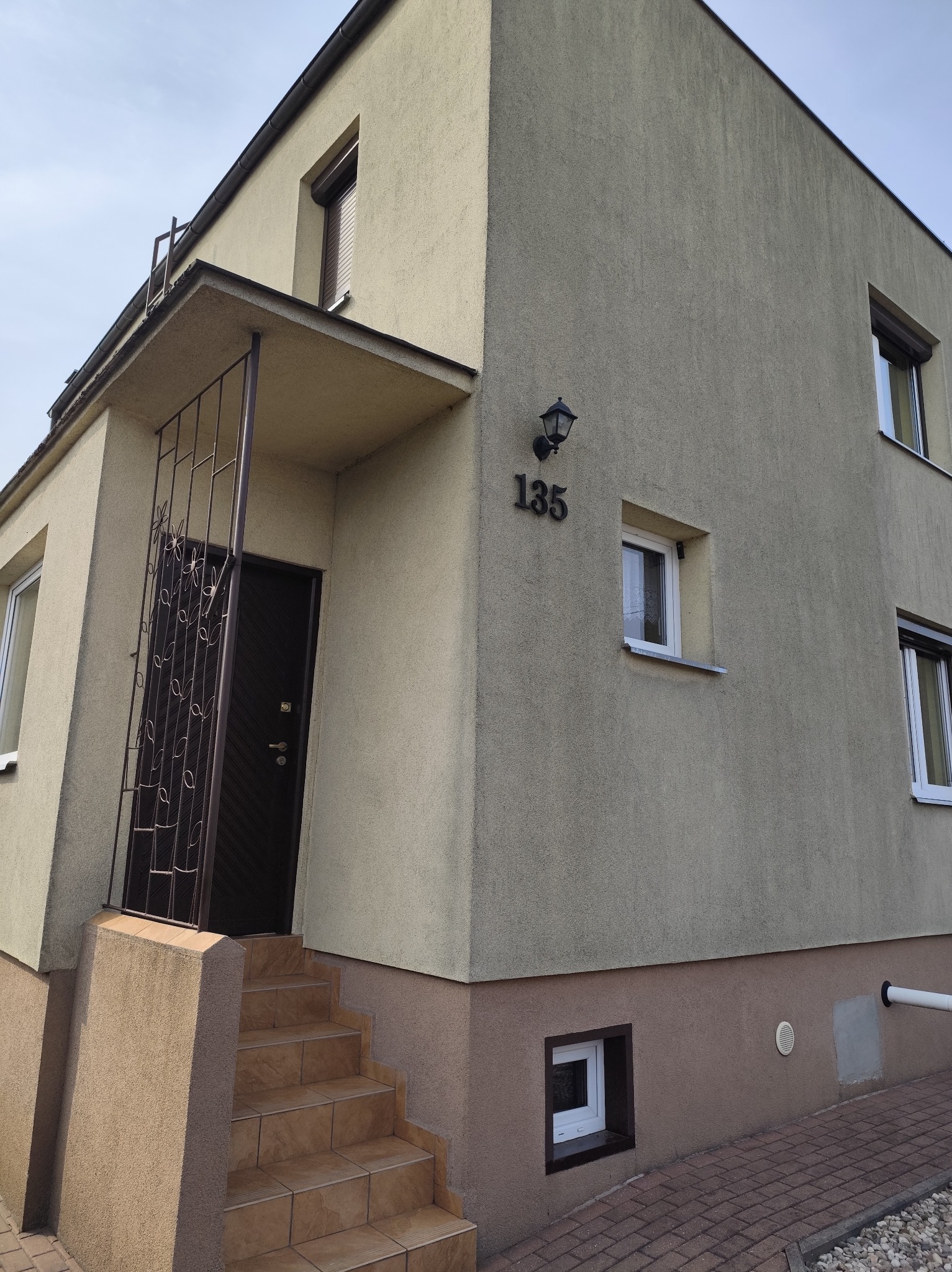 Dwukondygnacyjny dom z beżową elewacją i ciemnymi oknami. Wejście z metalową kratą i numerem 135. Widoczne schody wejściowe i fragment chodnika z kostki.