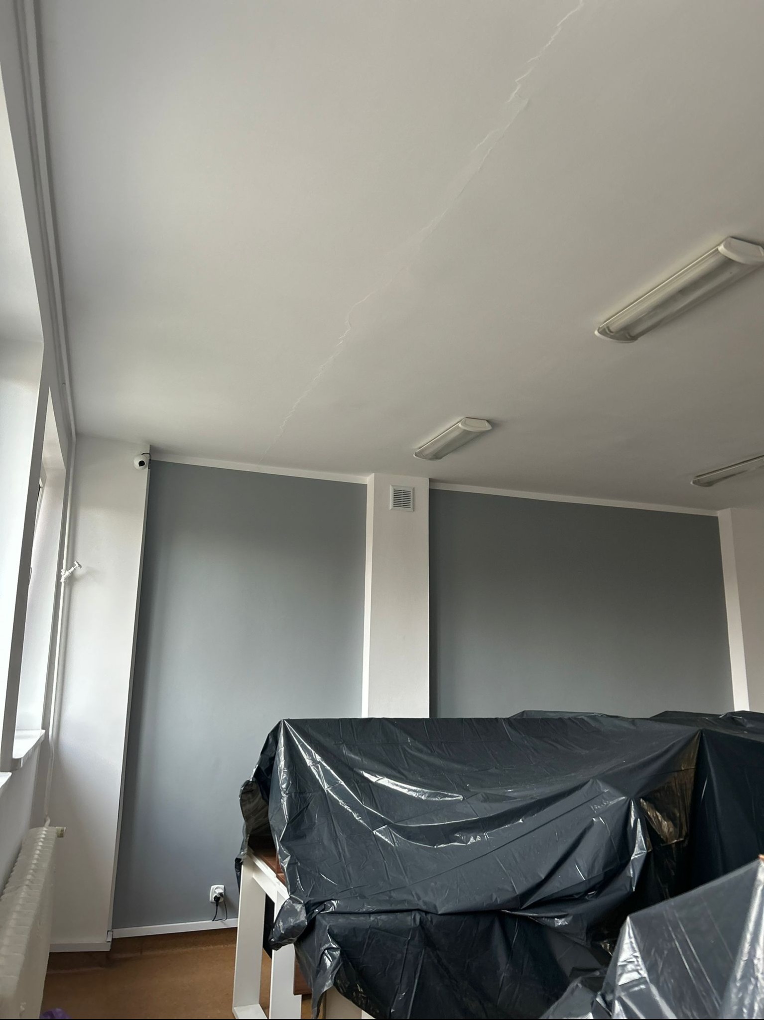 Przygotowanie do malowania: meble zabezpieczone folią, ściany pomalowane na szaro, sufit biały z widocznymi pęknięciami, oświetlenie jarzeniowe.