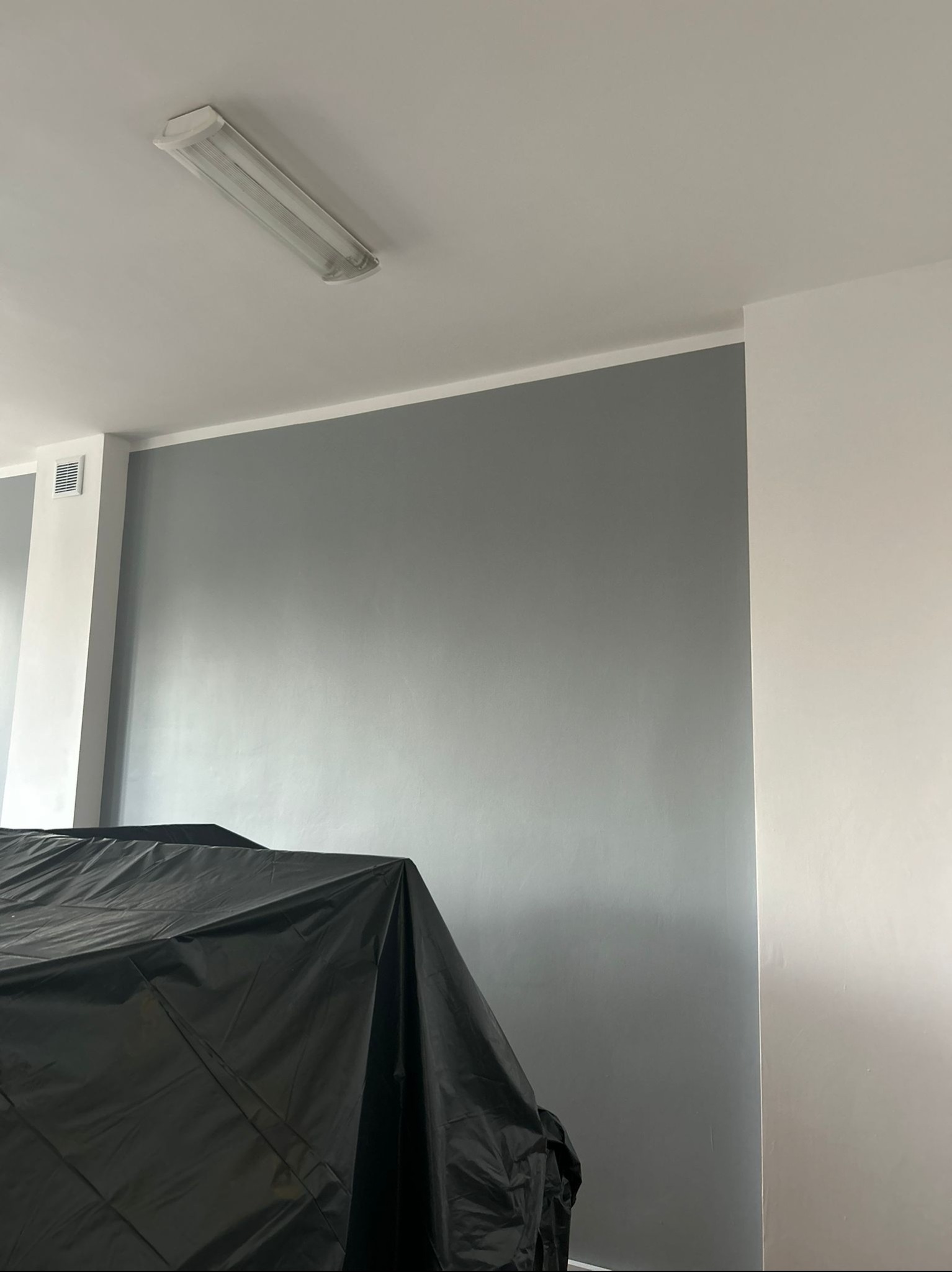 Ściana pomalowana na szaro, zabezpieczona czarną folią malarską. Widoczny fragment sufitu z lampą. Ujęcie z dołu, prezentujące etap prac malarskich.