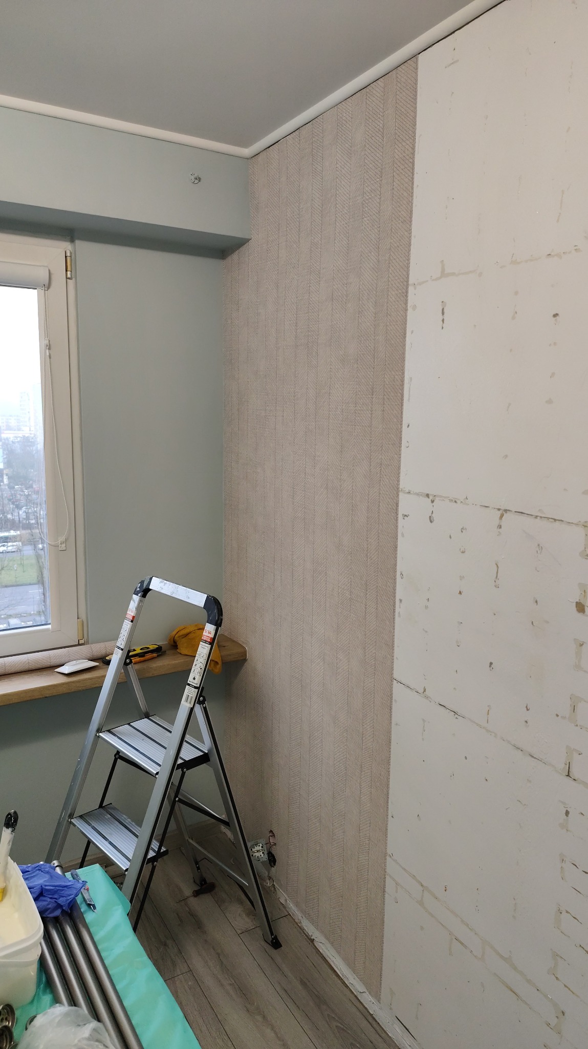 Ściana w trakcie tapetowania: fragment z wzorzystą tapetą, obok surowa ściana. Aluminiowa drabina i akcesoria malarskie przy oknie. Widok na miasto za oknem.