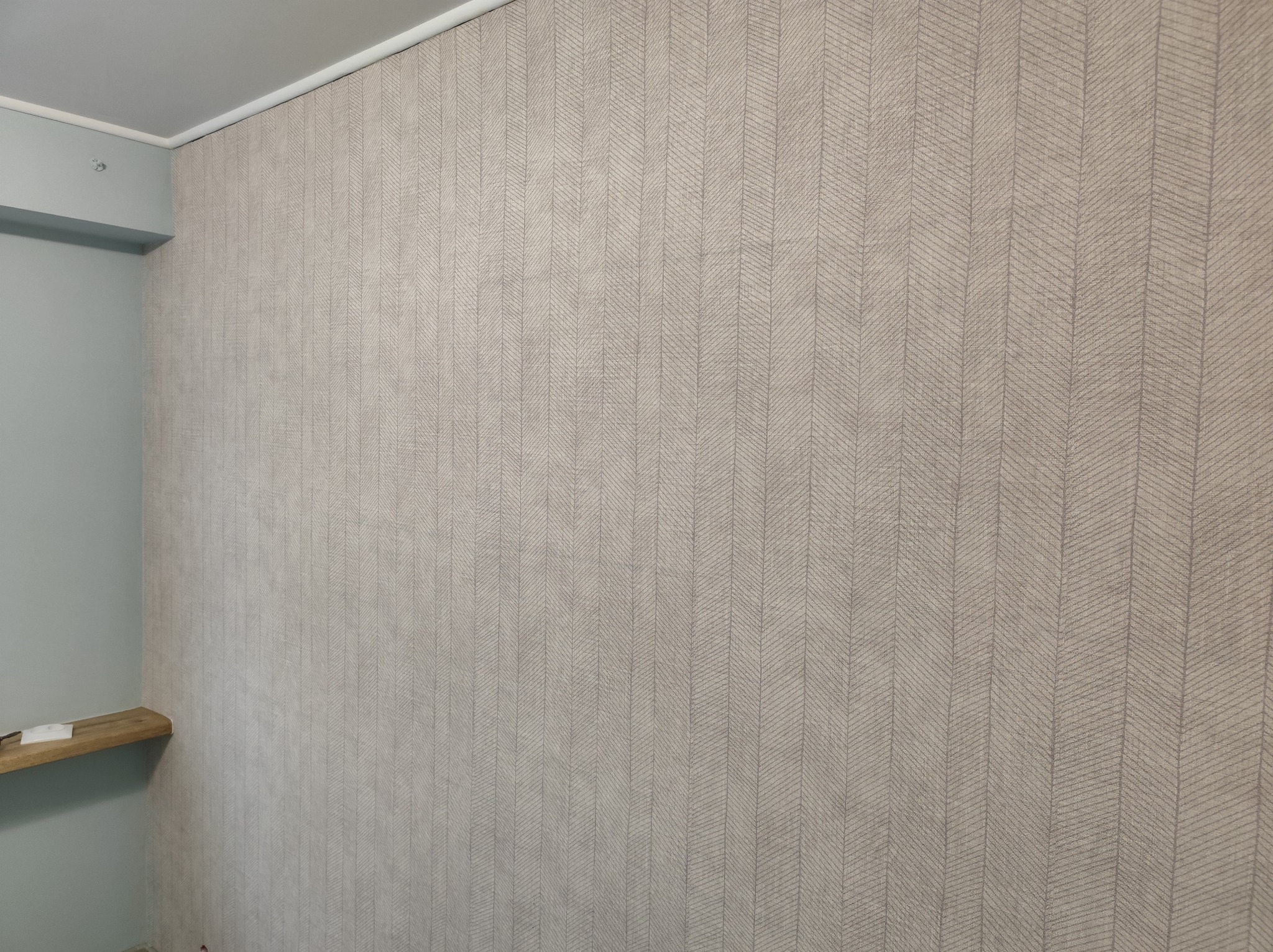 Ściana po malowaniu/tapetowaniu z wzorem w jodełkę, fragment półki i sufitu. Jasne, stonowane kolory. Widok na róg pokoju.