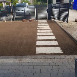 ZIELONA STREFA USŁUGI OGRODNICZE ANDRZEJ CHLEBIUK - Przygotowany teren pod trawnik z wytyczoną ścieżką z jasnych płyt. Na drugim planie czarne, nowoczesne ogrodzenie posesji i częściowo otwartą bramą wjazdową.