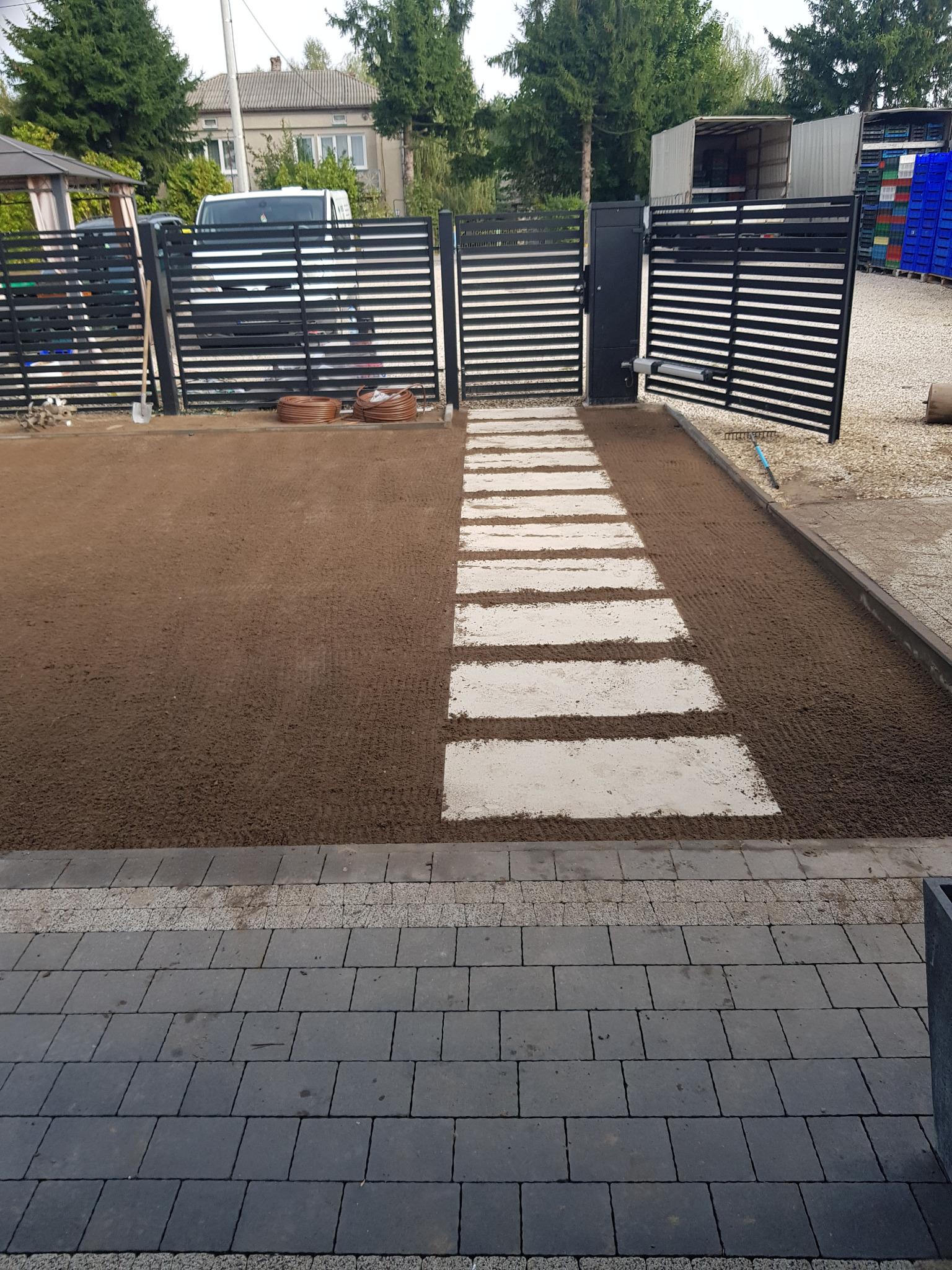 Przygotowany teren pod trawnik z wytyczoną ścieżką z jasnych płyt. Na drugim planie czarne, nowoczesne ogrodzenie posesji i częściowo otwartą bramą wjazdową.