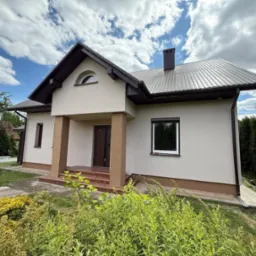 Dom jednorodzinny z jasną elewacją, brązowym dachem i stolarką okienną. Widoczne kolumny przy wejściu i zadbany ogród z bujną roślinnością. Słoneczny dzień.