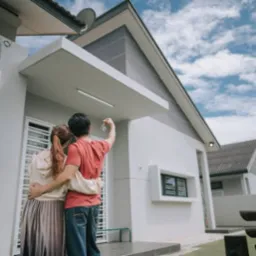 Para obejmuje się przed nowym, pomalowanym na biało domem z szarą elewacją szczytową. Mężczyzna trzyma klucze do domu w górze. Błękitne niebo z chmurami w tle.