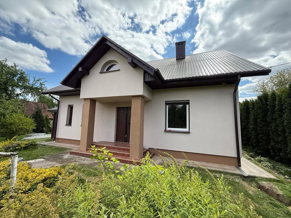 Dom jednorodzinny z jasną elewacją, brązowym dachem i stolarką okienną. Widoczne kolumny przy wejściu i zadbany ogród z bujną roślinnością. Słoneczny dzień.