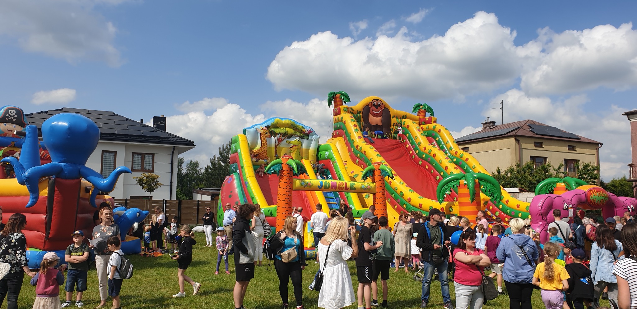 Kolorowy, dmuchany plac zabaw z motywem dżungli, zjeżdżalnie i postacie zwierząt, otoczony przez bawiące się dzieci i dorosłych w słoneczny dzień.
