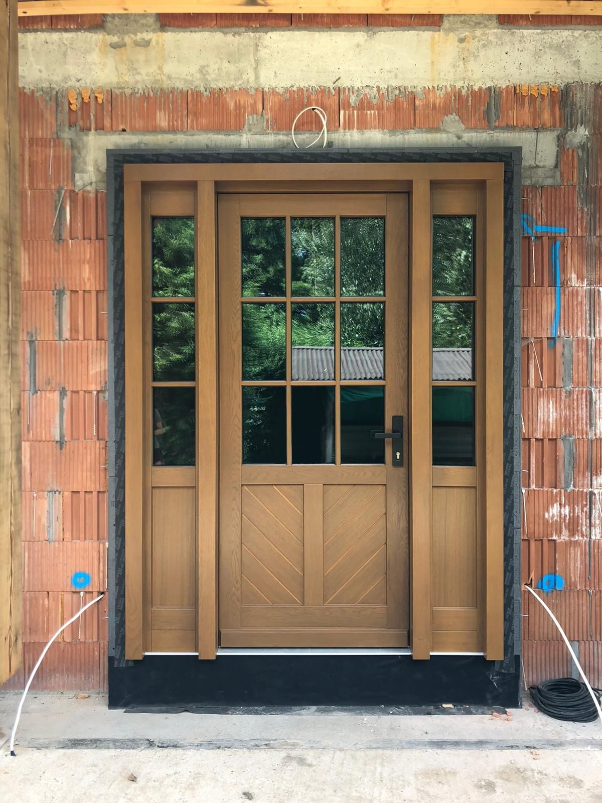 Drewniane drzwi wejściowe z szybami, zamontowane w surowym murze z czerwonej cegły. Widoczne kable i instalacje, co sugeruje etap wykończeniowy.