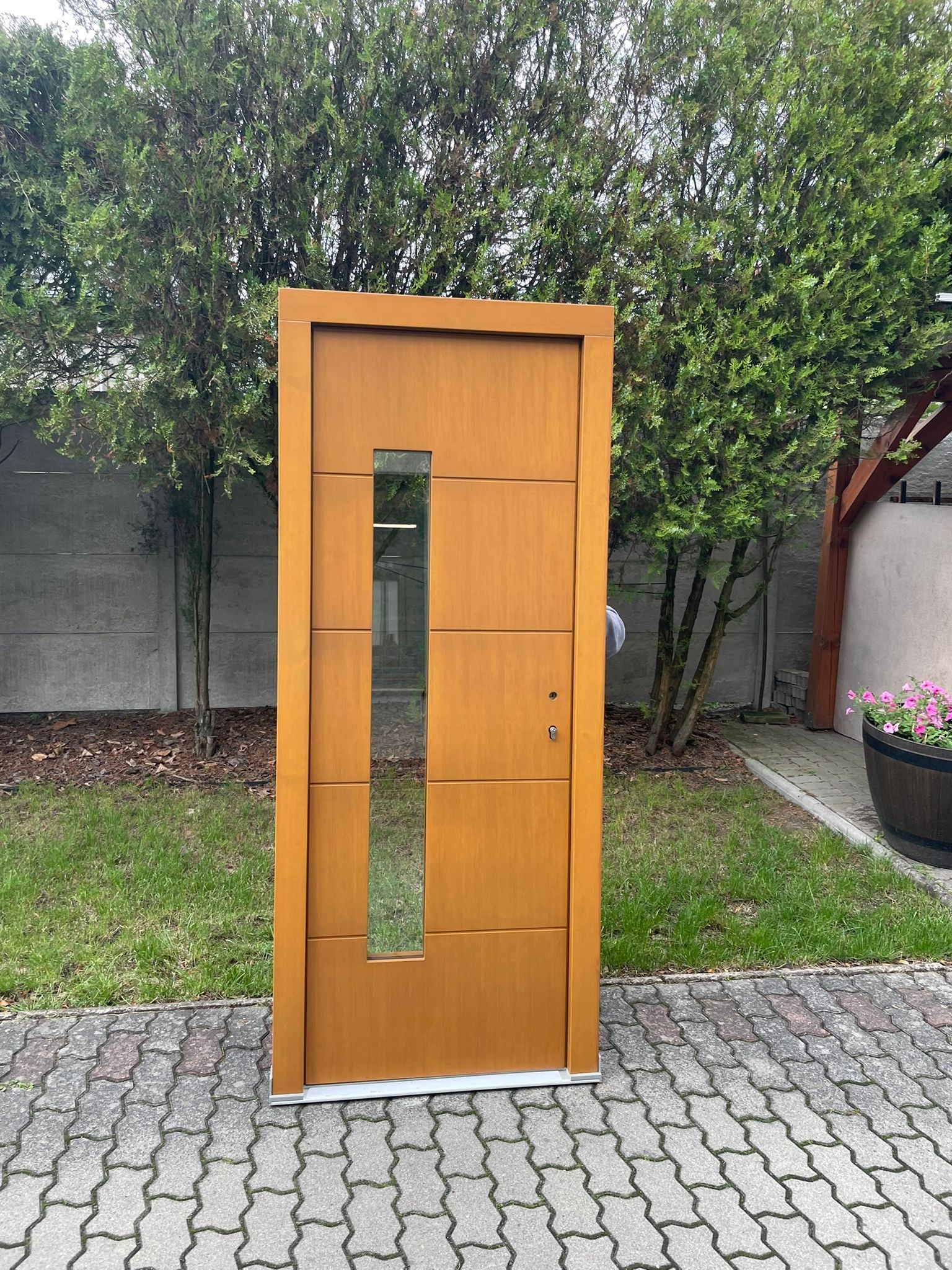 Złote drzwi wejściowe z szybą, ustawione na brukowanym podjeździe, otoczone zielenią drzew i trawy, z widocznym elementem architektury ogrodowej w tle.