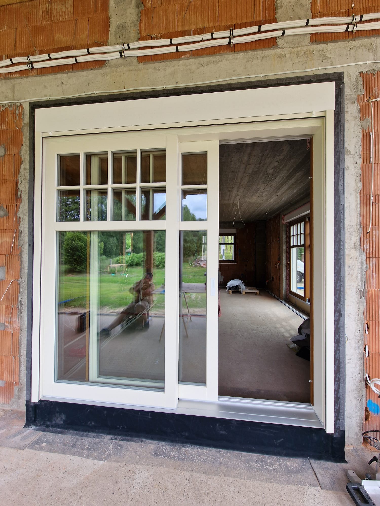 Białe okno przesuwne HS z roletą zewnętrzną, widok z zewnątrz na wnętrze surowego budynku z ceglanymi ścianami. Częściowo otwarte, z widocznym odbiciem osoby.