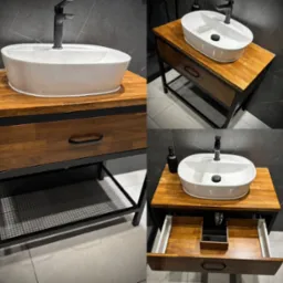 Nowoczesna umywalka nablatowa owalna na szafce z drewnianym blatem i czarną metalową ramą, widoczne otwarte szuflady i półka z metalowej siatki, czarna bateria, szara płytka na ścianie i podłodze.