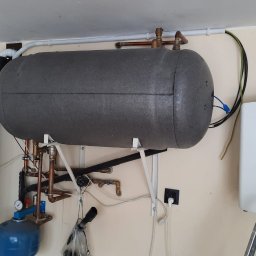 TRANSMAR MARCIN KĘPSKI - Szary, izolowany zbiornik akumulacyjny ciepła zawieszony na ścianie, z widocznymi rurami miedzianymi, obok biały inwerter solarny Solis i szary odkurzacz centralny Beam z niebieskim podświetleniem,...