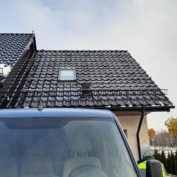 TRANSMAR MARCIN KĘPSKI - Czarny dach z dachówki ceramicznej z zamontowanymi szynami montażowymi pod instalację paneli słonecznych, widok zza niebieskiego samochodu dostawczego z odbiciami w szybie.