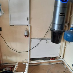TRANSMAR MARCIN KĘPSKI - Szary odkurzacz centralny BEAM zamontowany na beżowej ścianie, obok instalacja hydrauliczna z niebieskim zbiornikiem, podłączony do instalacji elektrycznej widocznej na ścianie, na podłodze...