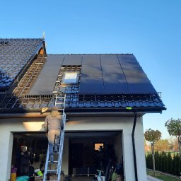 TRANSMAR MARCIN KĘPSKI - Pracownik na drabinie montuje panele słoneczne na dachu domu z dachówką ceramiczną, widoczne okno dachowe i częściowo zamontowane panele, garaż z otwartymi drzwiami w tle.