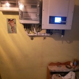 TRANSMAR MARCIN KĘPSKI - Biała skrzynka z podłączonymi przewodami i wyświetlaczem, zamontowana na żółtej ścianie w pomieszczeniu, obok skrzynka z bezpiecznikami i rurki instalacyjne.