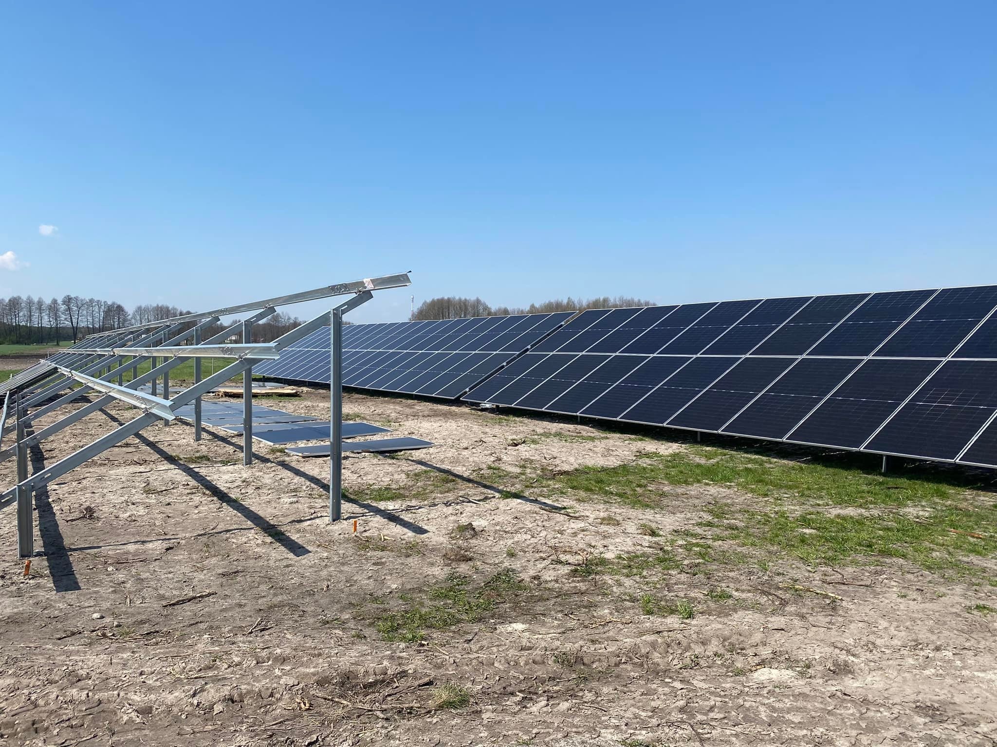 Rząd paneli słonecznych w trakcie montażu na polu, z widoczną metalową konstrukcją wsporczą i niezamontowanymi panelami obok.