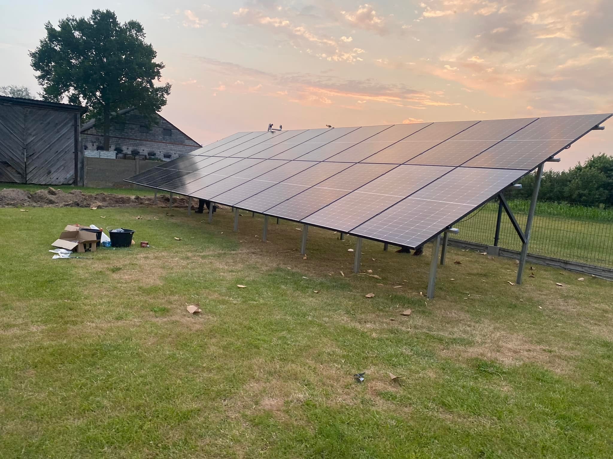 Panele słoneczne na trawniku w trakcie montażu, widoczne narzędzia i kartony, w tle budynek gospodarczy i zachodzące słońce.