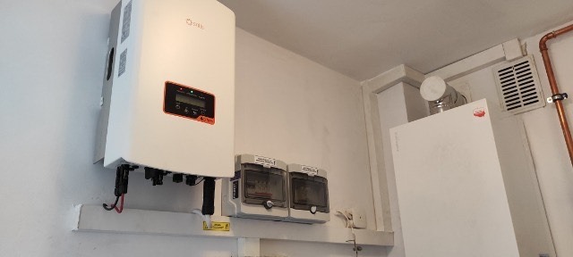 Biały inwerter fotowoltaiczny marki Solis zamontowany na ścianie obok dwóch szarych skrzynek bezpiecznikowych i białego pieca centralnego ogrzewania z miedzianymi rurami.