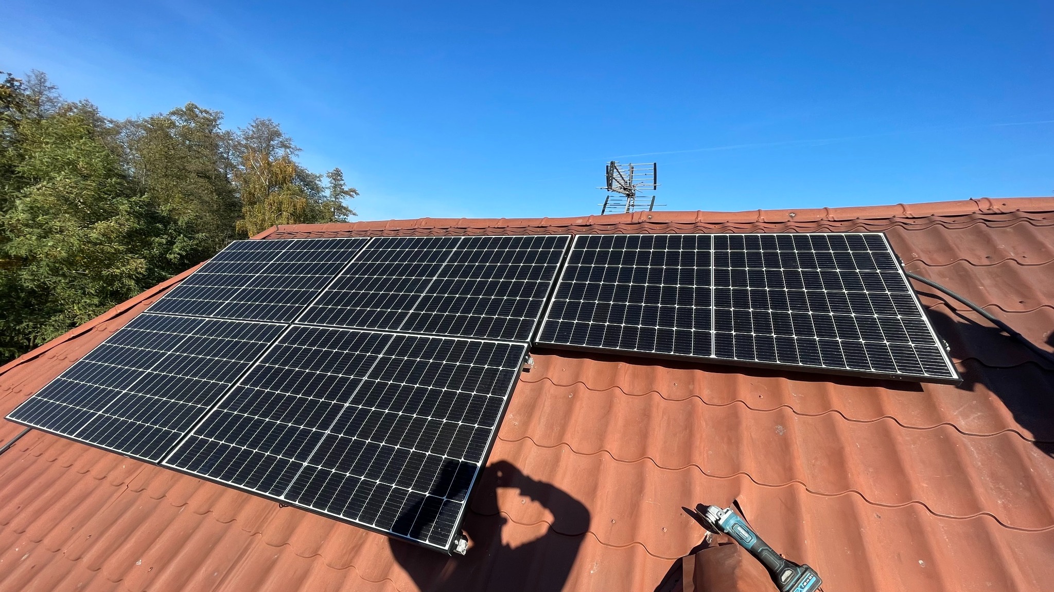 Panele słoneczne na czerwonym dachu z widoczną anteną telewizyjną w tle, cień osoby i szlifierka kątowa sugerują trwający montaż.