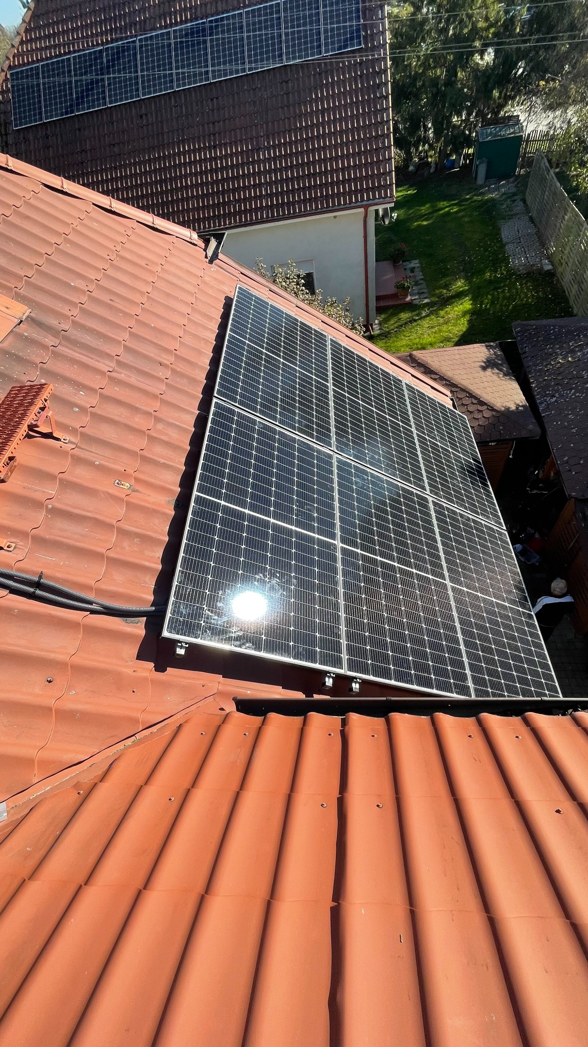 Panel fotowoltaiczny zamontowany na dachu pokrytym czerwoną dachówką, widoczne czarne kable prowadzące wzdłuż dachu, odbicie słońca na powierzchni paneli.