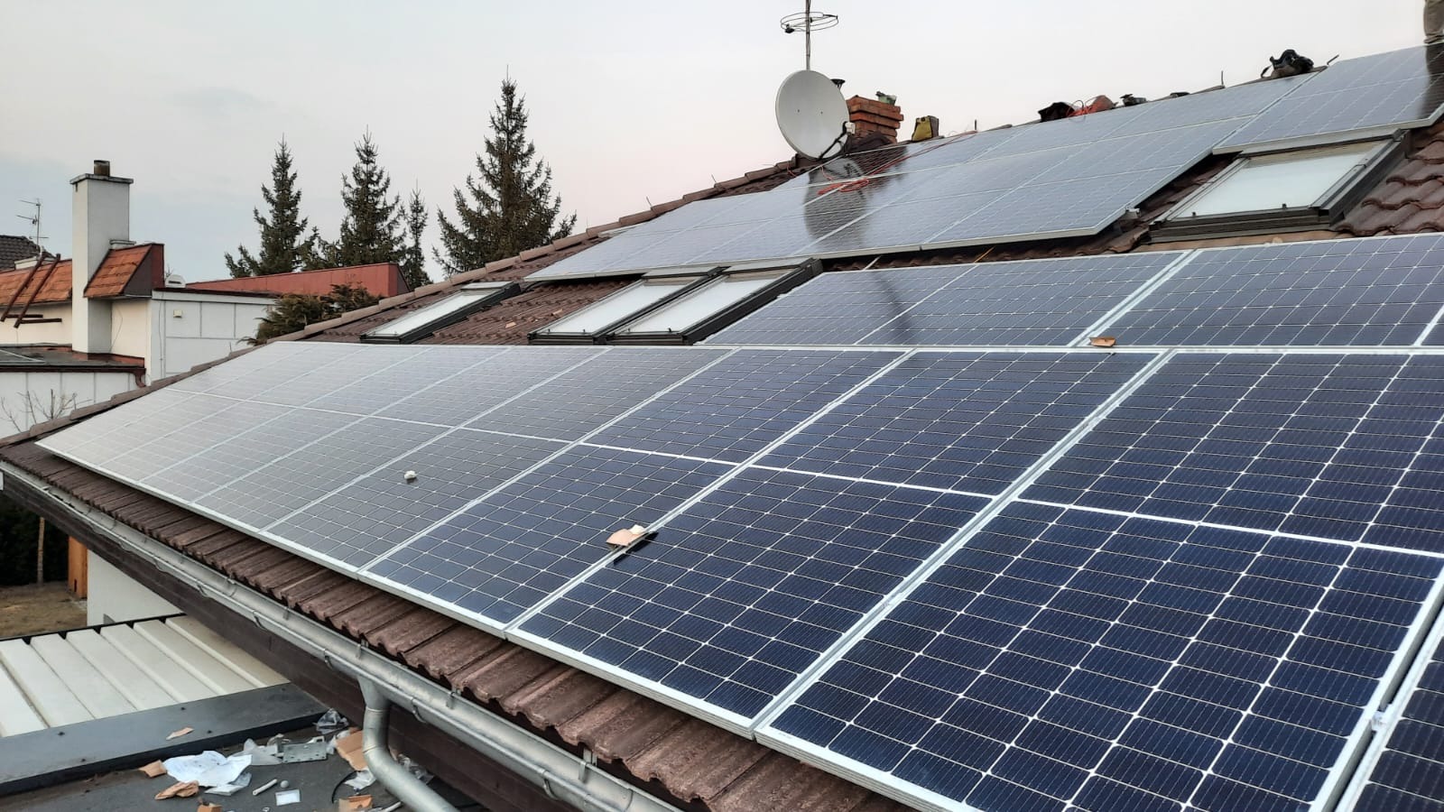 Panele słoneczne na dachu domu z brązową dachówką, widoczne okna dachowe i antena satelitarna, na jednym z paneli leży liść.