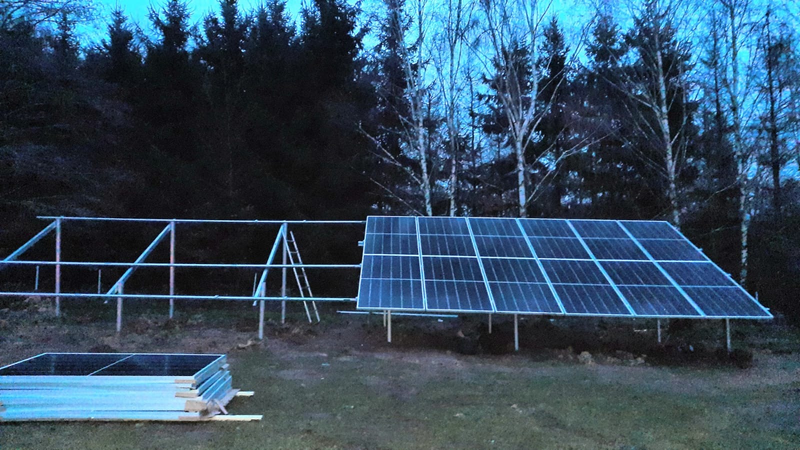 Panel słoneczny zamontowany na metalowej konstrukcji w plenerze, w tle widoczne drzewa, obok stos paneli przygotowanych do montażu.