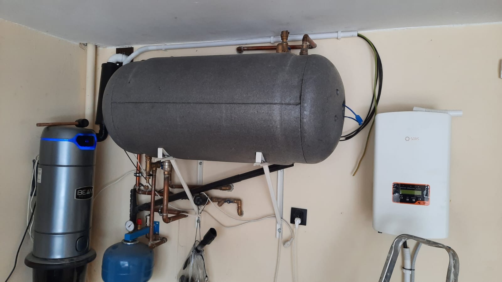 Szary, izolowany zbiornik akumulacyjny ciepła zawieszony na ścianie, z widocznymi rurami miedzianymi, obok biały inwerter solarny Solis i szary odkurzacz centralny Beam z niebieskim podświetleniem,...