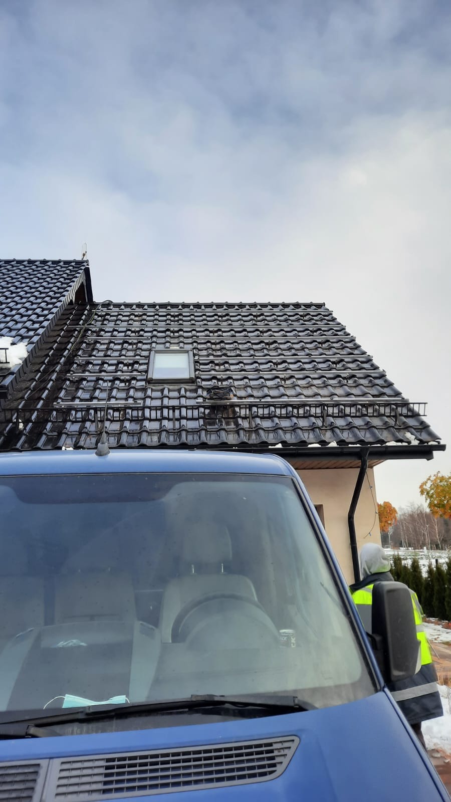 Czarny dach z dachówki ceramicznej z zamontowanymi szynami montażowymi pod instalację paneli słonecznych, widok zza niebieskiego samochodu dostawczego z odbiciami w szybie.