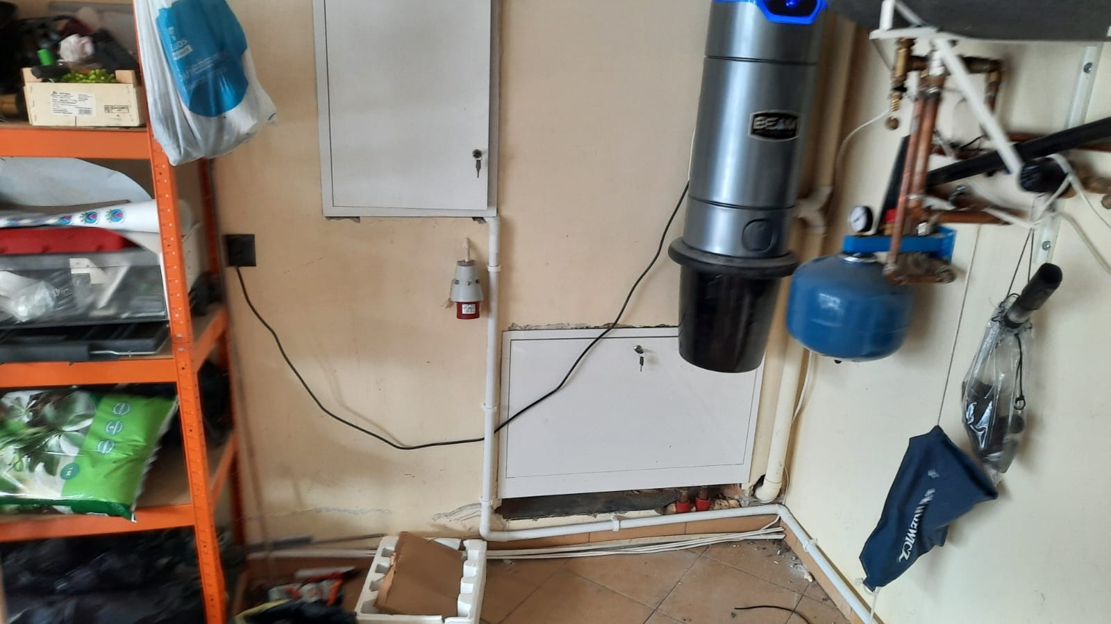 Szary odkurzacz centralny BEAM zamontowany na beżowej ścianie, obok instalacja hydrauliczna z niebieskim zbiornikiem, podłączony do instalacji elektrycznej widocznej na ścianie, na podłodze...