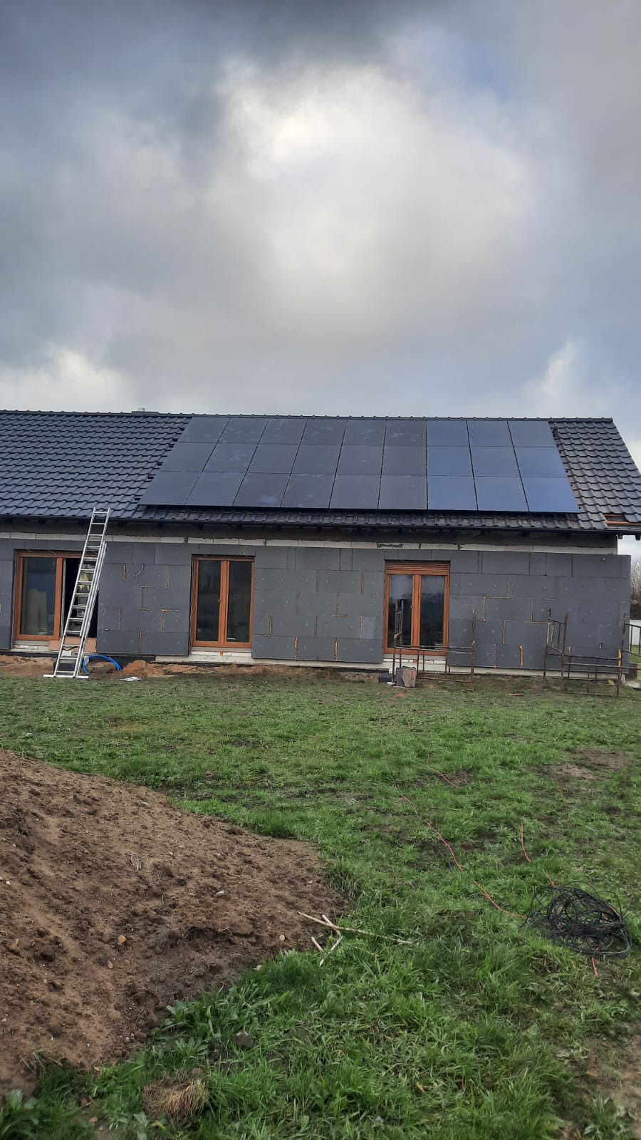 Dom jednorodzinny w trakcie montażu paneli słonecznych na dachu, widoczna drabina oparta o ścianę budynku, ocieplenie ścian styropianem grafitowym, pochmurne niebo.