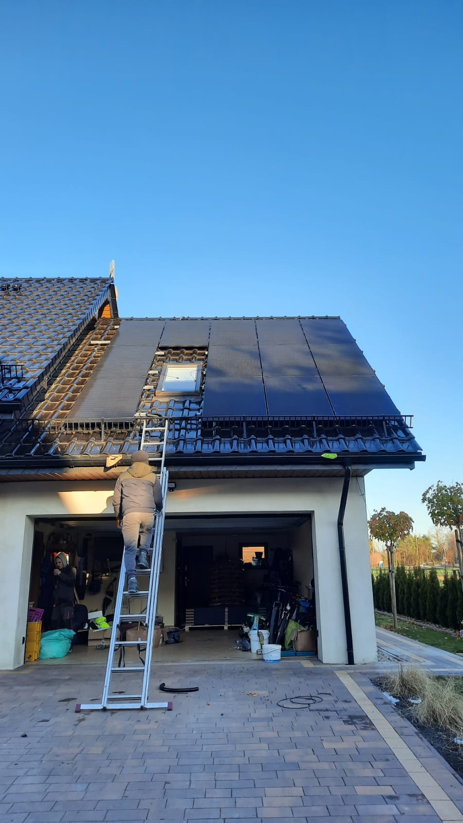 Pracownik na drabinie montuje panele słoneczne na dachu domu z dachówką ceramiczną, widoczne okno dachowe i częściowo zamontowane panele, garaż z otwartymi drzwiami w tle.