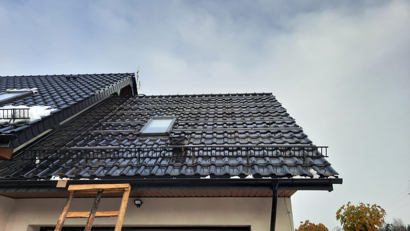 Częściowo zamontowane, metalowe szyny na dachu pokrytym ciemną dachówką, przygotowane pod instalację paneli słonecznych, widoczne okno dachowe i drabina oparta o budynek.