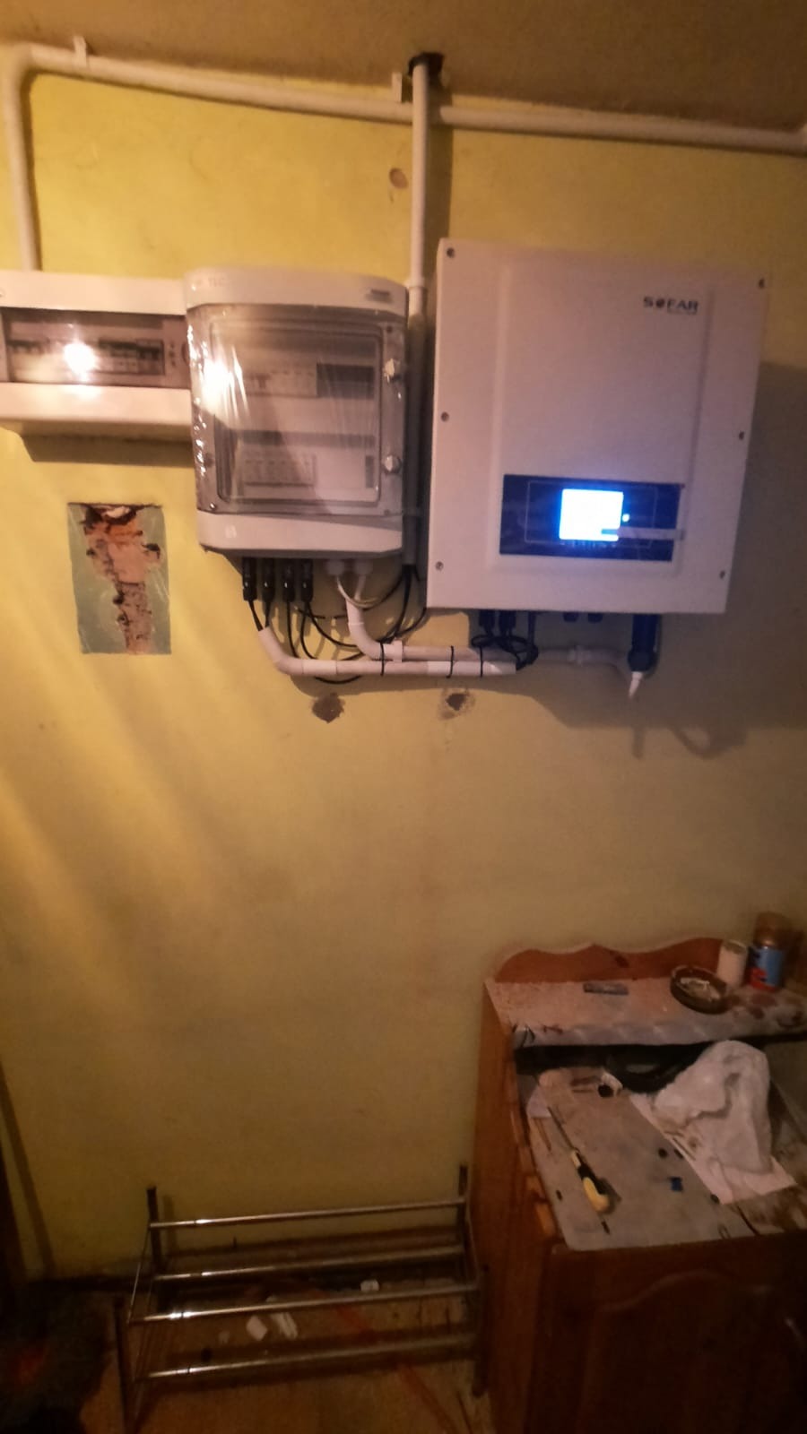 Biała skrzynka z podłączonymi przewodami i wyświetlaczem, zamontowana na żółtej ścianie w pomieszczeniu, obok skrzynka z bezpiecznikami i rurki instalacyjne.