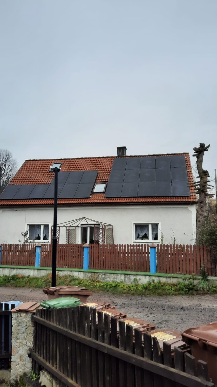 Dom jednorodzinny z panelami słonecznymi na dachu z czerwonej dachówki, widok z poziomu ulicy w pochmurny dzień. Na pierwszym planie widoczny fragment drewnianego ogrodzenia pomalowanego na brązowo...