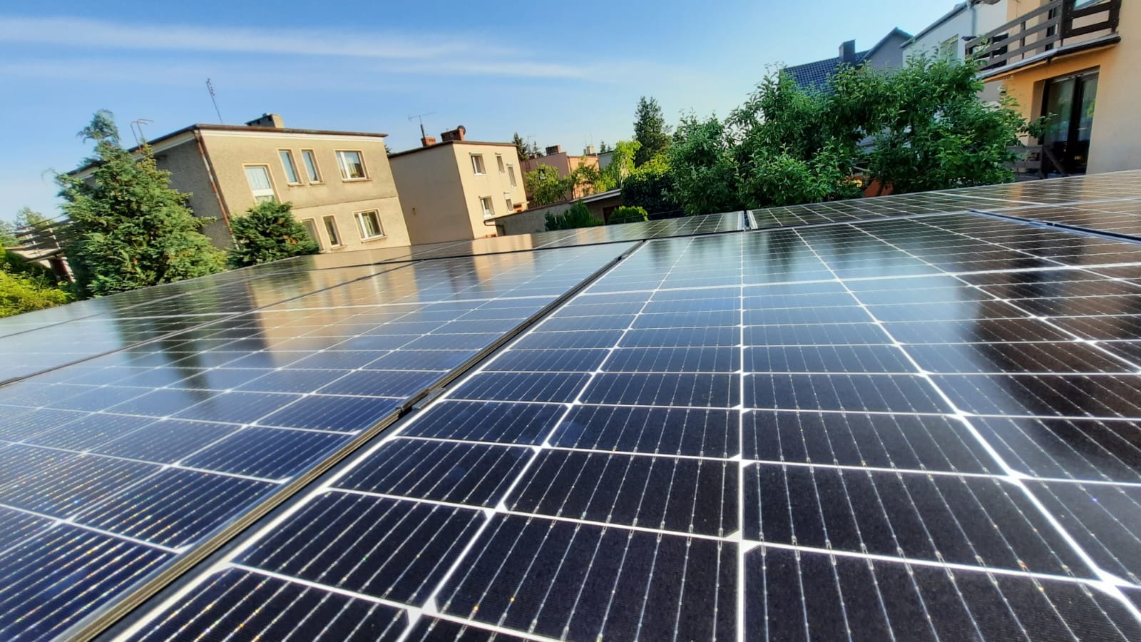 Błyszczące panele słoneczne na dachu budynku mieszkalnego, odbijające niebo i otaczającą zieleń, widziane z perspektywy lekko z dołu.