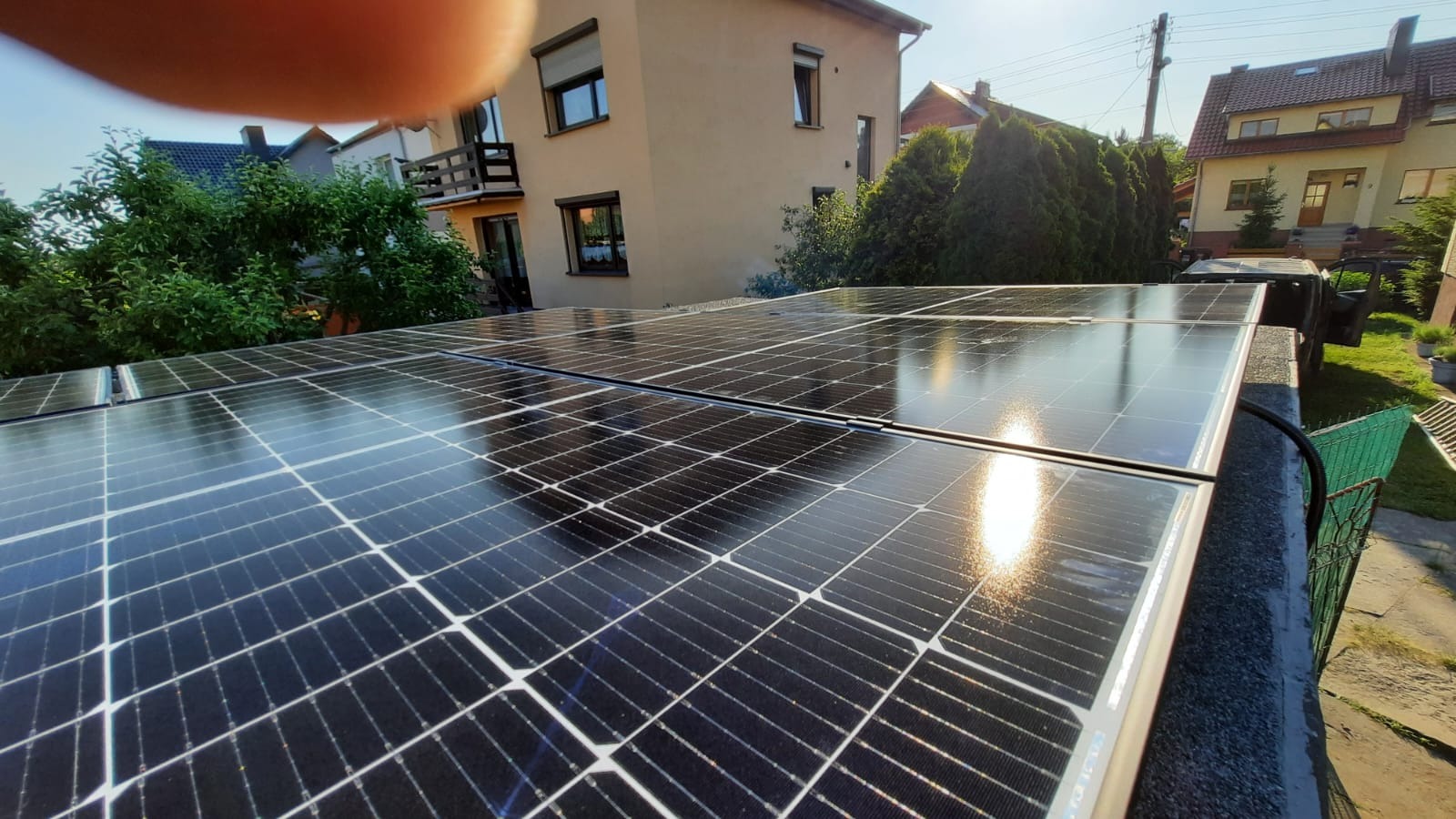 Błyszczące panele fotowoltaiczne na dachu z widocznym odbiciem słońca, na tle zabudowań mieszkalnych i zieleni.