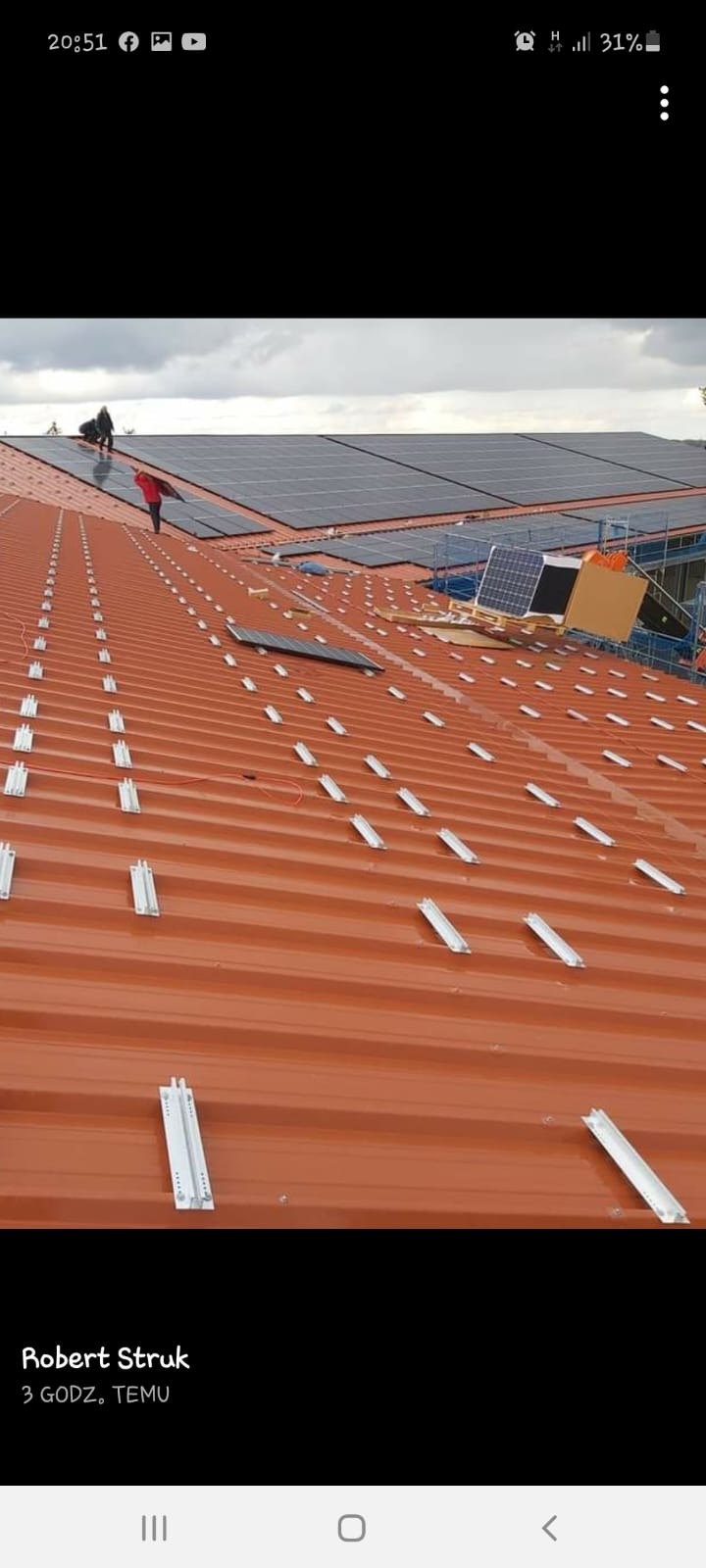Montaż paneli na dachu z blachy trapezowej, widoczne szyny montażowe, pracownicy na dachu instalują panele w pochmurny dzień.