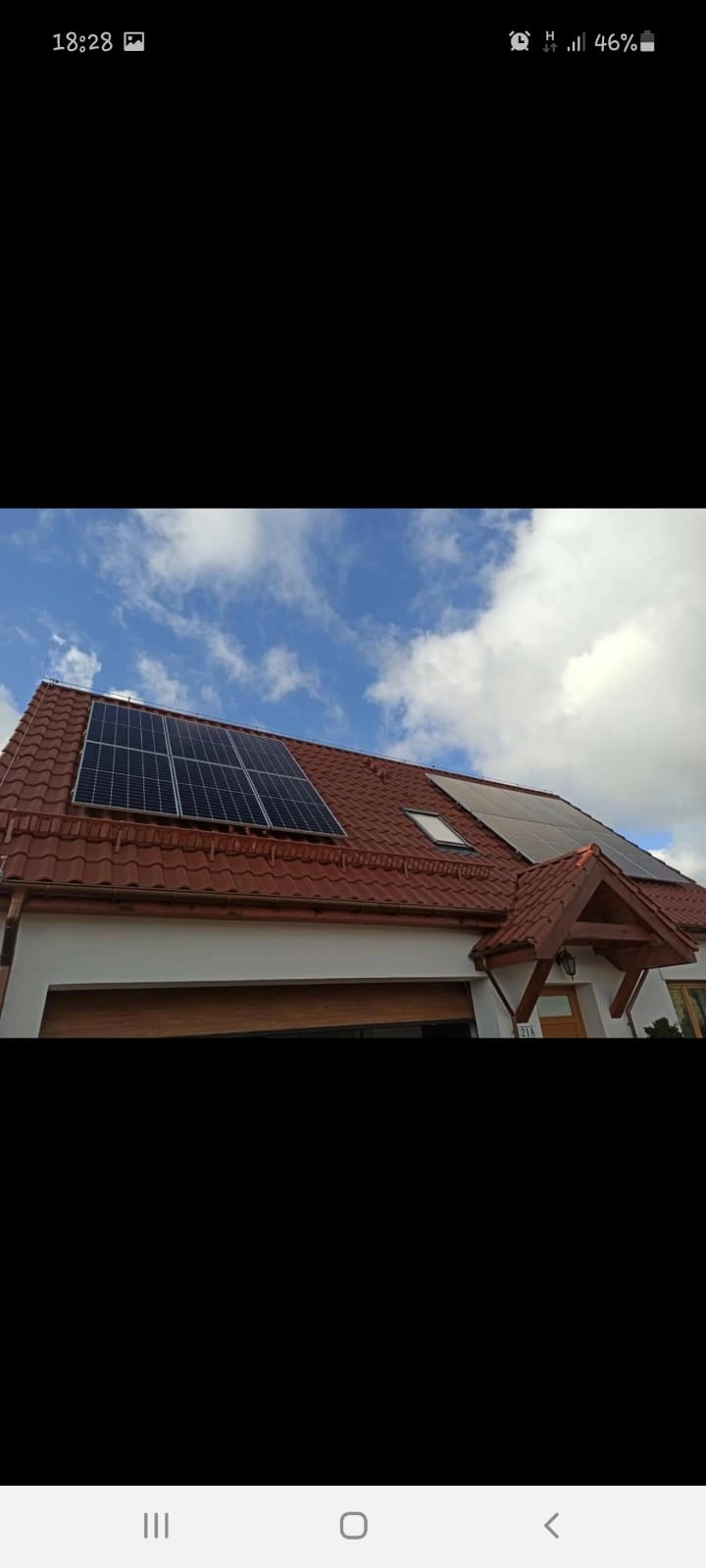 Dom jednorodzinny z czerwoną dachówką i zamontowanymi panelami słonecznymi na dachu, widoczne okno dachowe i błękitne niebo z chmurami w tle.